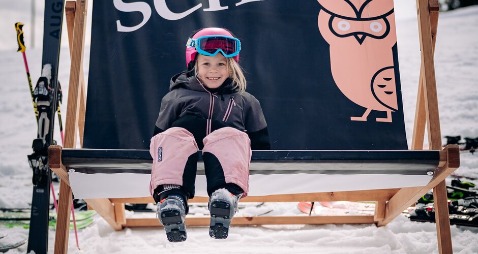 Lächelndes Kind mit Skibekleidung sitzt im Schnee auf großem Liegestuhl mit Banner Rose im Schnee