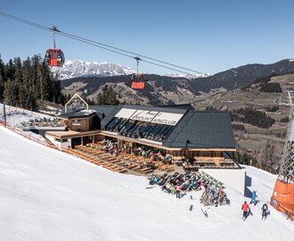 Mountain lodge with sun terrace on a ski slope, red gondola cabins above and snow-covered peaks behind