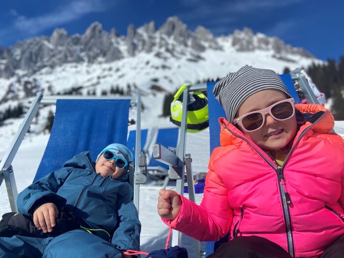 Two children relaxing in lounge chairs with sunglasses in front of snowy mountains. | © Anna Pilz