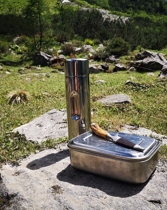 Metal lunchbox, knife and thermos bottle placed on rock in alpine meadow
