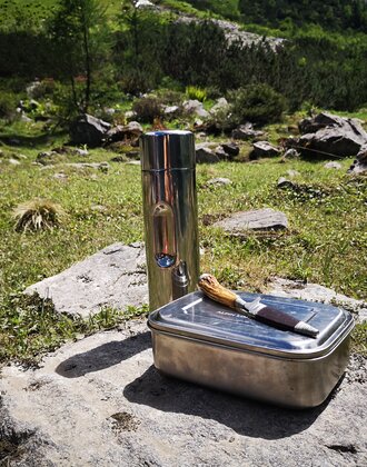 Metal lunchbox, knife and thermos bottle placed on rock in alpine meadow