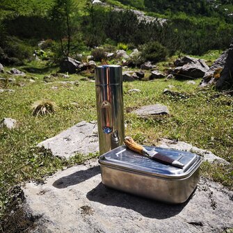 Metal lunchbox, knife and thermos bottle placed on rock in alpine meadow