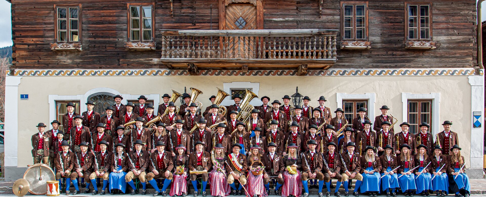 The Altenmarkt traditional band poses in costume with instruments in front of a rustic wooden house | © Trachtenmusikkapelle Altenmarkt