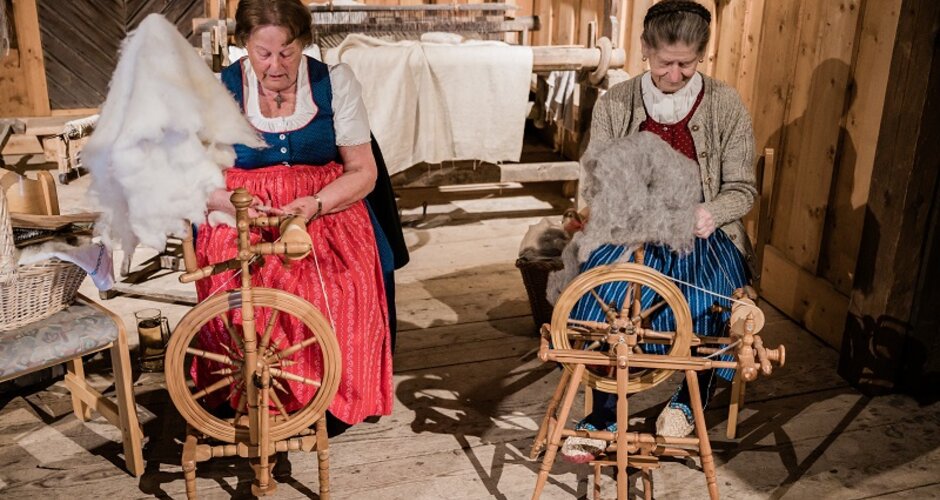 Zwei Frauen in Tracht spinnen Schafwolle am Spinnrad in einer alten Stube beim Handwerks-Event in traditionellem Ambiente. | © Matthias Fritzenwallner