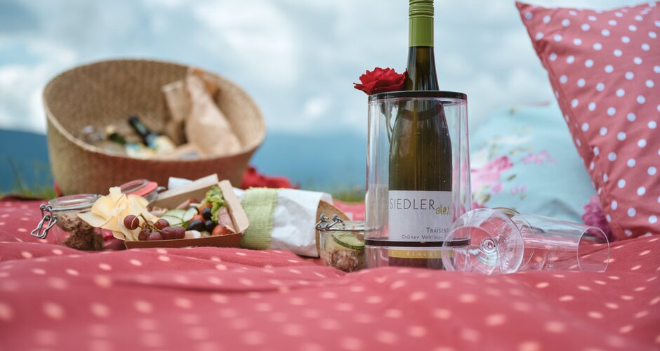 Wine bottle, glasses and picnic snacks on a dotted blanket at the See.Musik event, with mountains and sky in the background | © Matthias Fritzenwallner