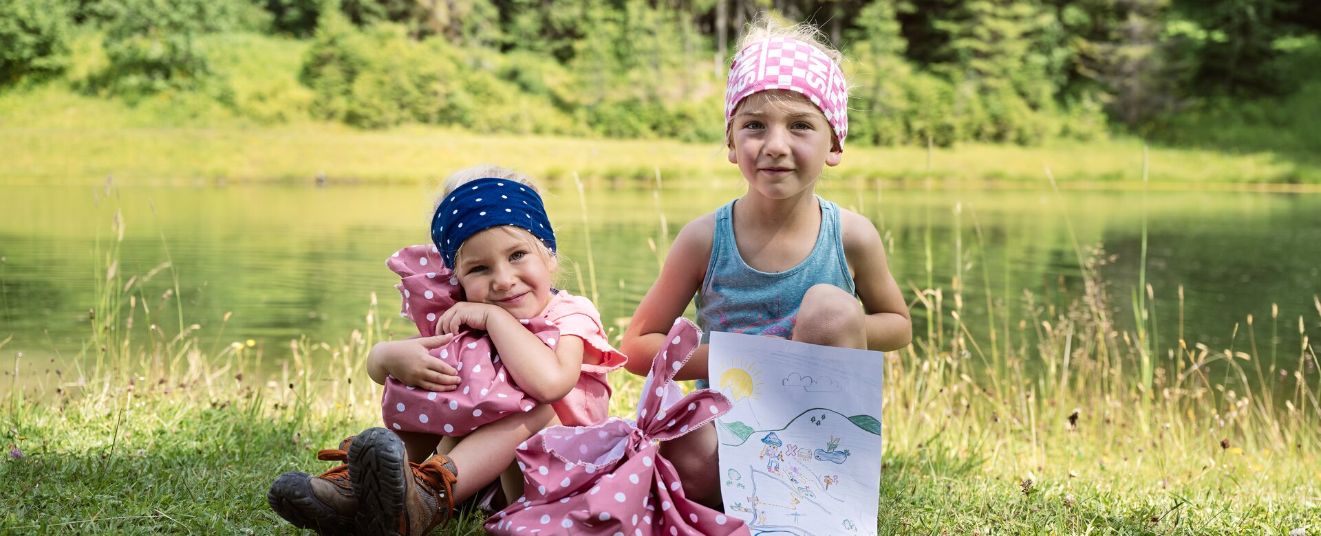 Two kids at the See.Musik event by the lake, sitting on grass with picnic bags and a self-drawn picture of sun and mountains. | © Matthias Fritzenwallner