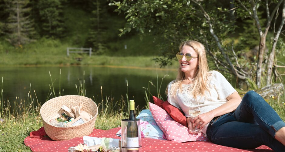 Woman relaxing on a dotted blanket with wine and picnic basket by the lake during the See.Musik event in the sunshine | © Matthias Fritzenwallner