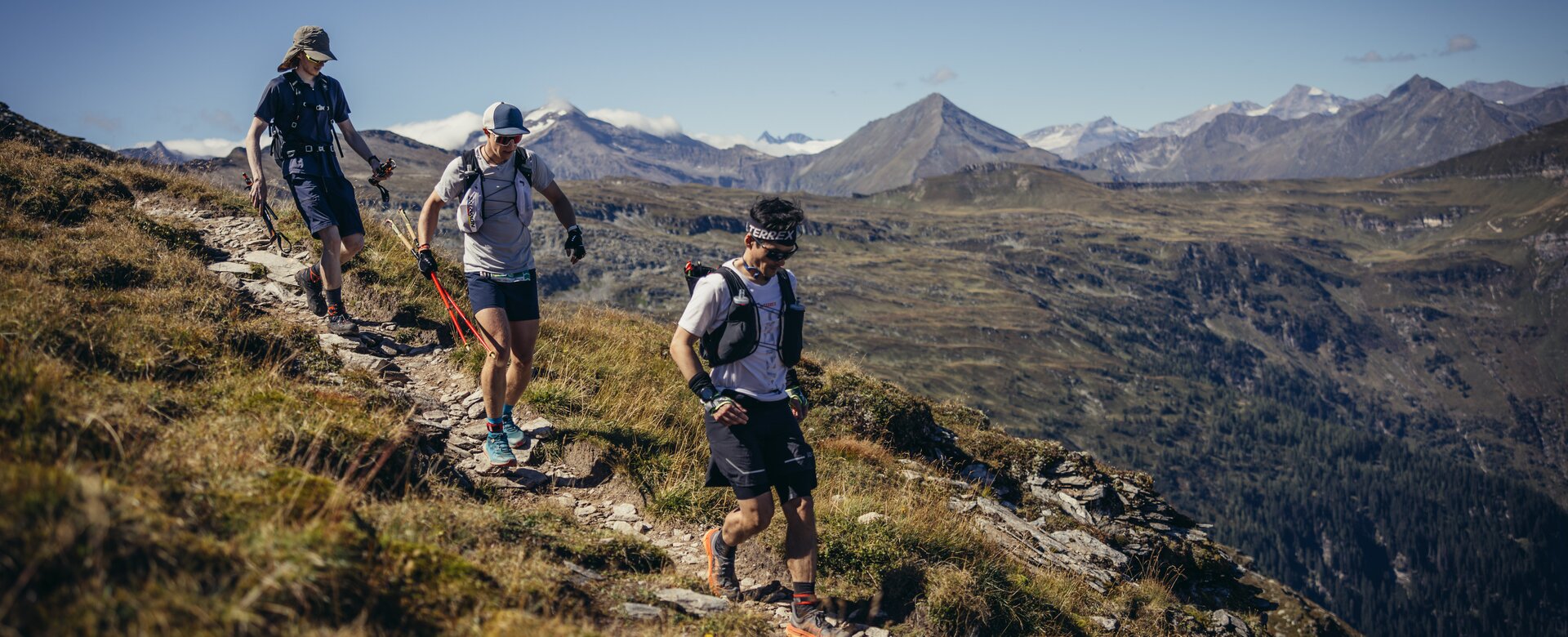 Three trail runners hiking along a rocky mountain path with green valleys and snowy High Tauern peaks in the background | © Stefan Schaaf PEAK ART IMAGES