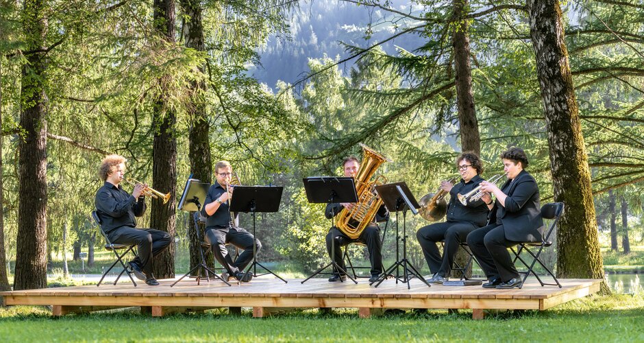 A brass ensemble performs on a forest stage, surrounded by trees and summer light in Gastein | © Gasteinertal Tourismus GmbH, Simon Hutter