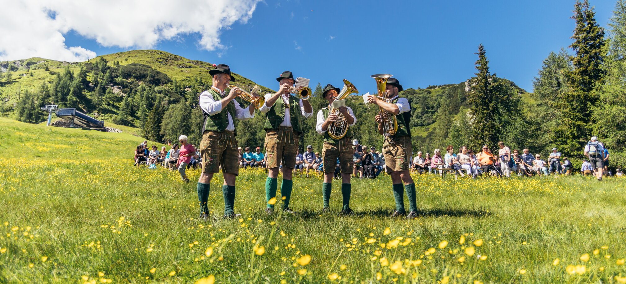 4 musicians are standing on a meadow playing brass instruments and many people are sitting in the background | © Gerald Grünwald
