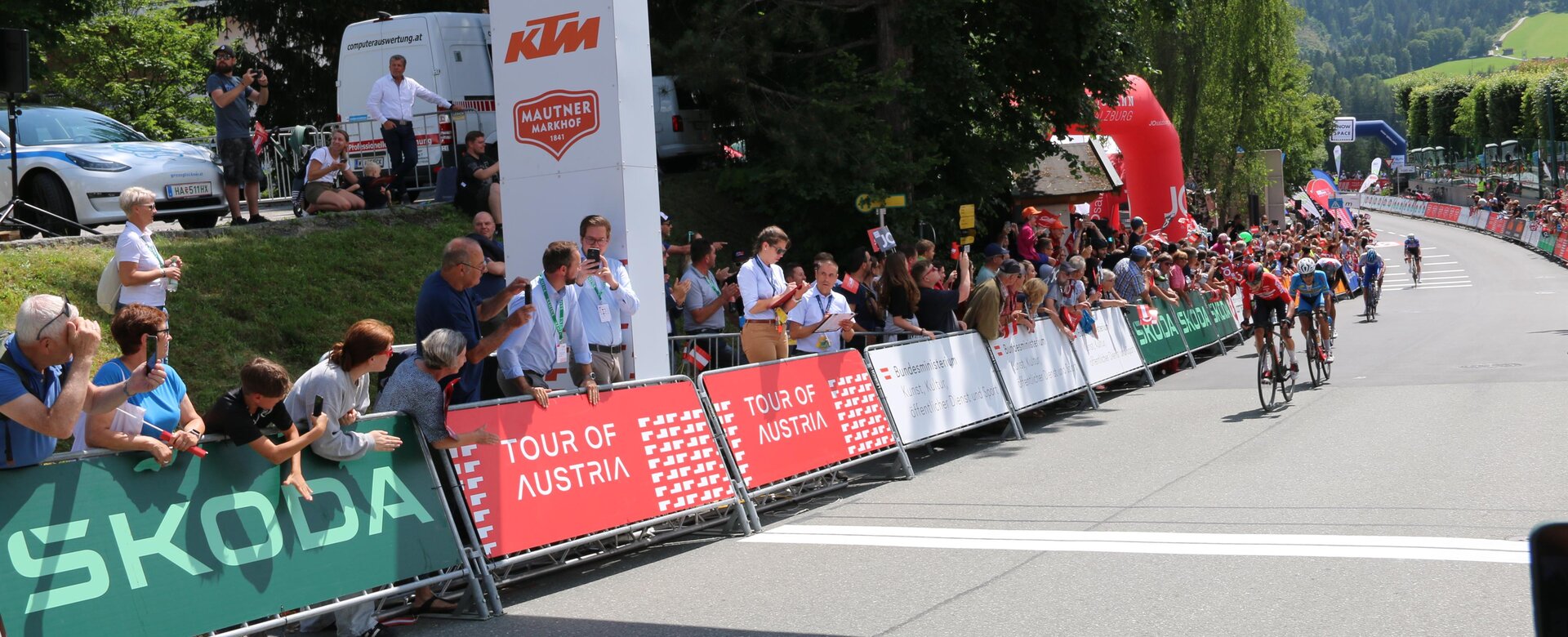 Cyclists cross the finish line at the Tour of Austria as spectators cheer by the roadside and finish arch | © JOSalzburg