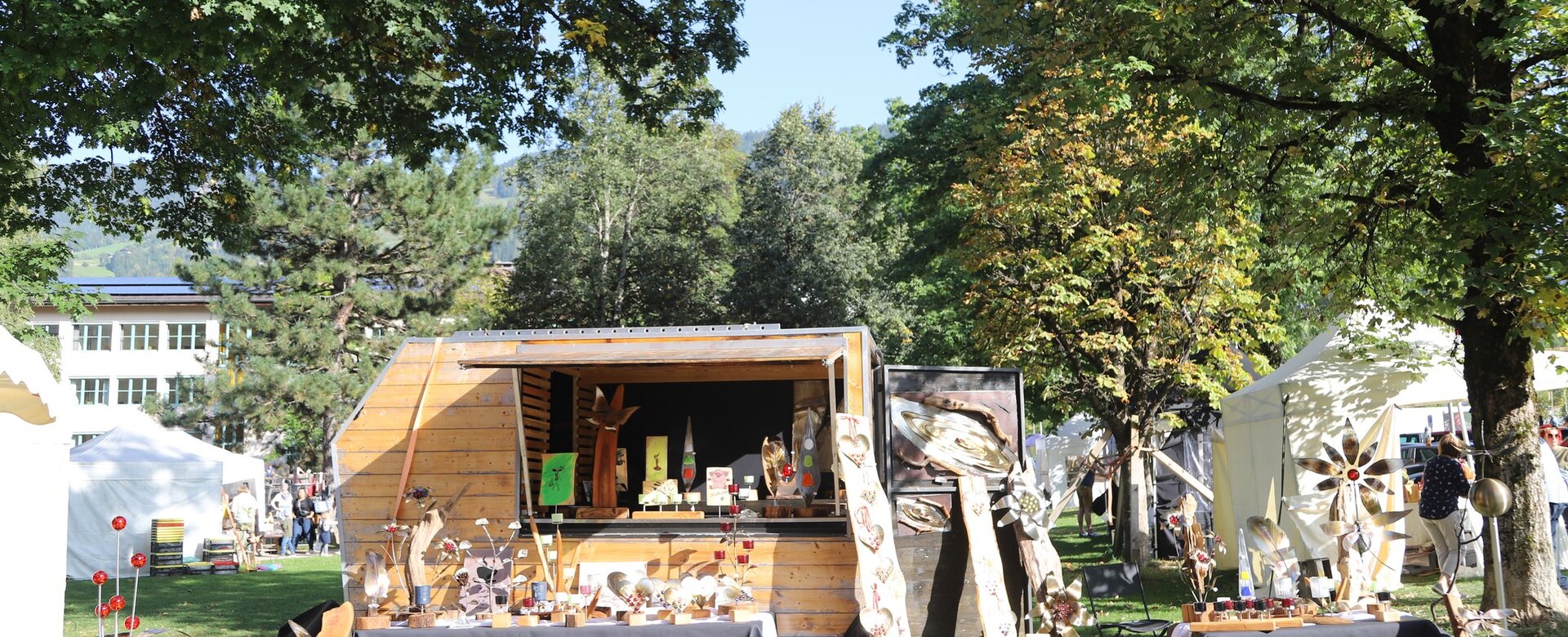Market stall with wood art, metal decor and crafts in park, sunny day, white tents in background | © JOSalzburg