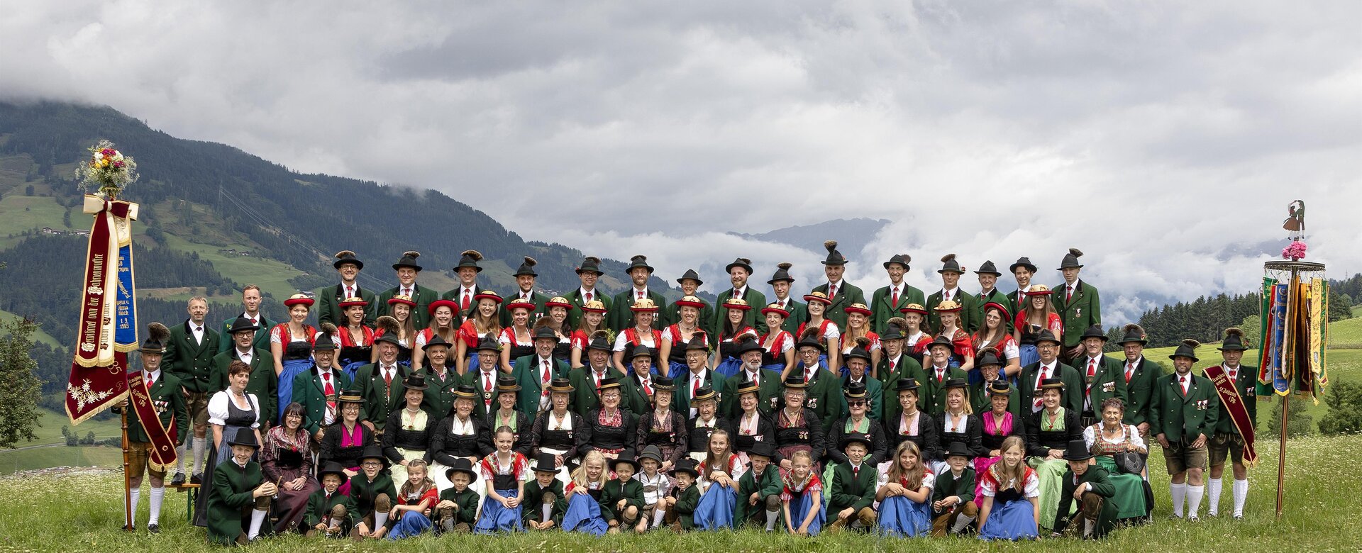 Large traditional group with two banners on alpine meadow, men in green, women in dirndl, children in front | © Trachtenverein