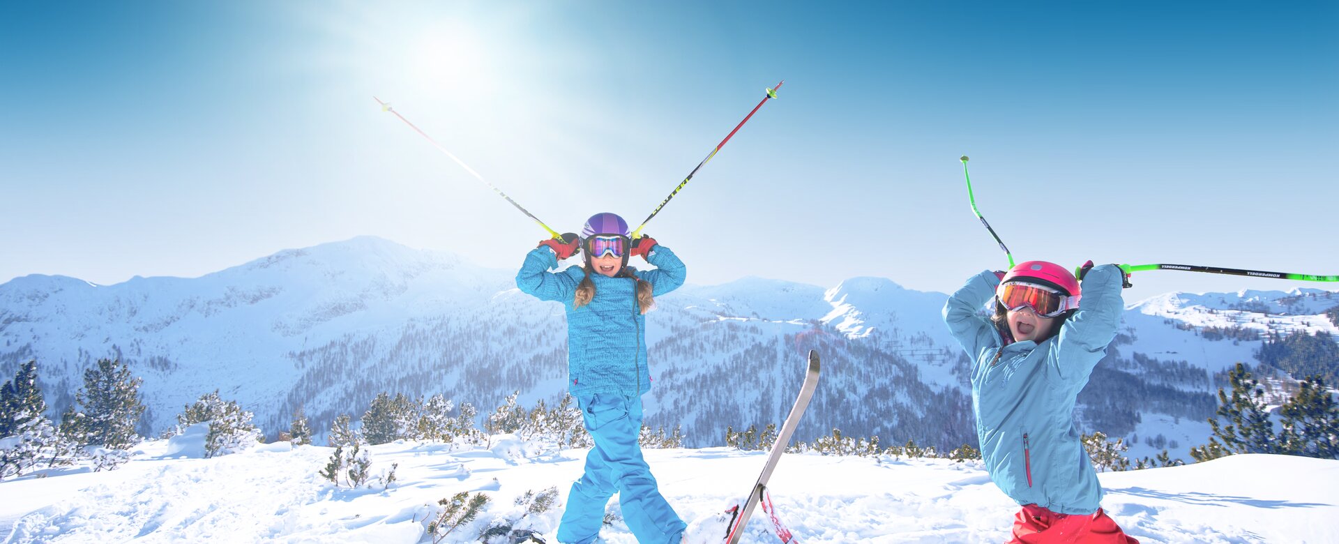 Two children are standing in deep snow, holding their ski poles to their helmets so that they look like horns, with their mouths wide open