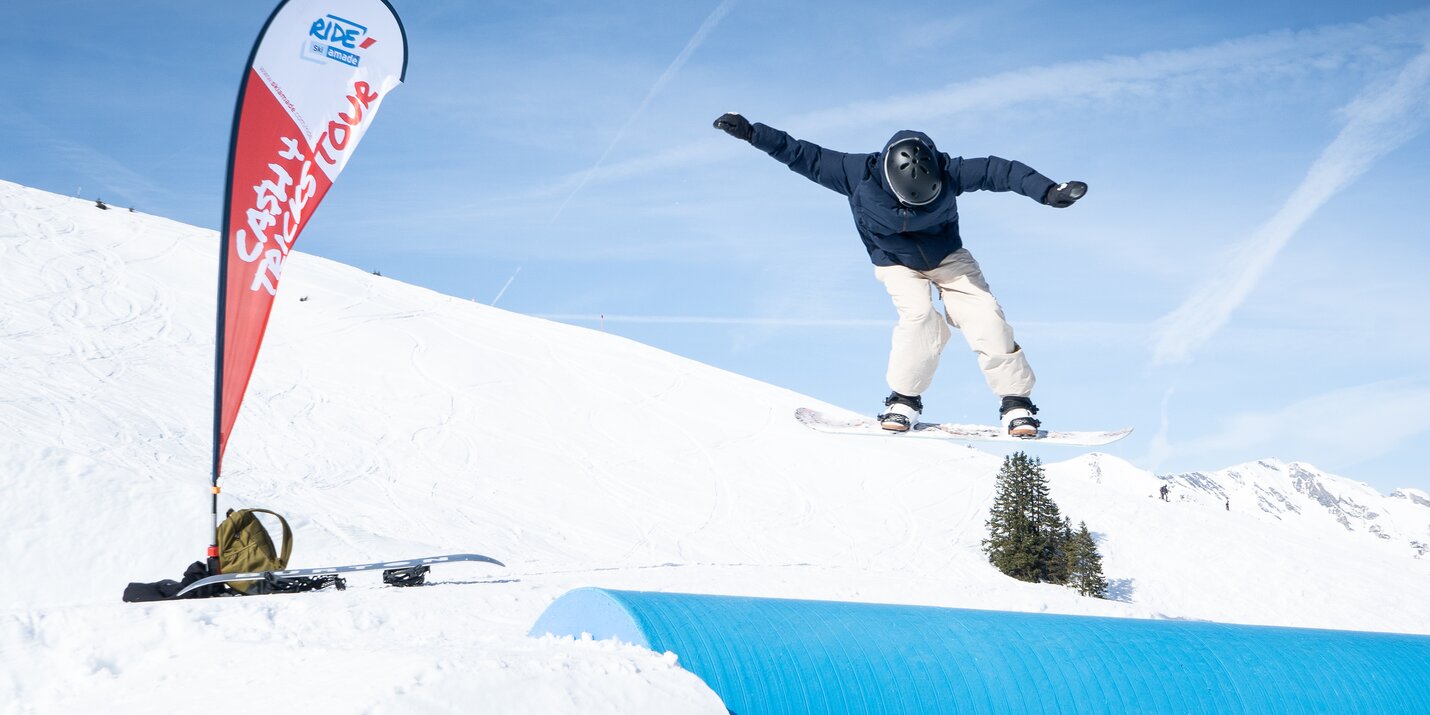 Snowboarder jumps over blue tube feature at Cash4Tricks with clear blue sky, red event flag and gear placed on the snow nearby.