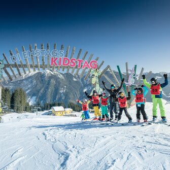Kindergruppe mit Skiern posiert jubelnd unter dem „Kidstag“-Bogen im Skigebiet Shuttleberg | © Shuttleberg