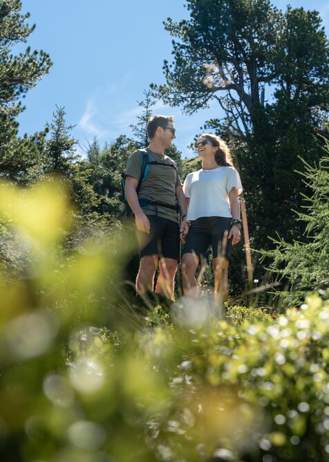 Smiling couple hiking through forest at Graukogel in sunshine, surrounded by alpine greenery | © Skigastein, seven frames