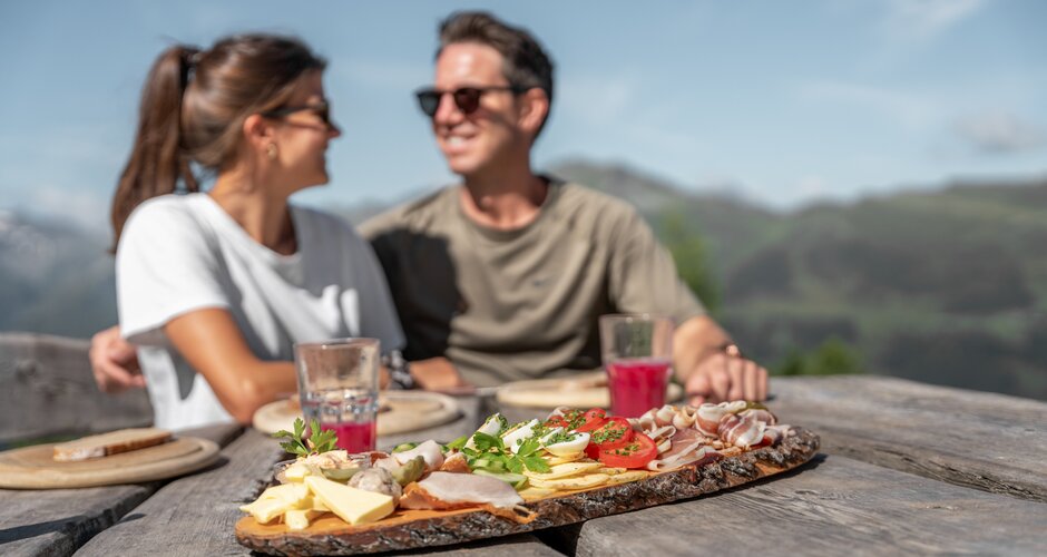 Couple enjoys alpine snack with cheese, bacon, juice on wooden table and mountain view | © Skigastein, seven frames