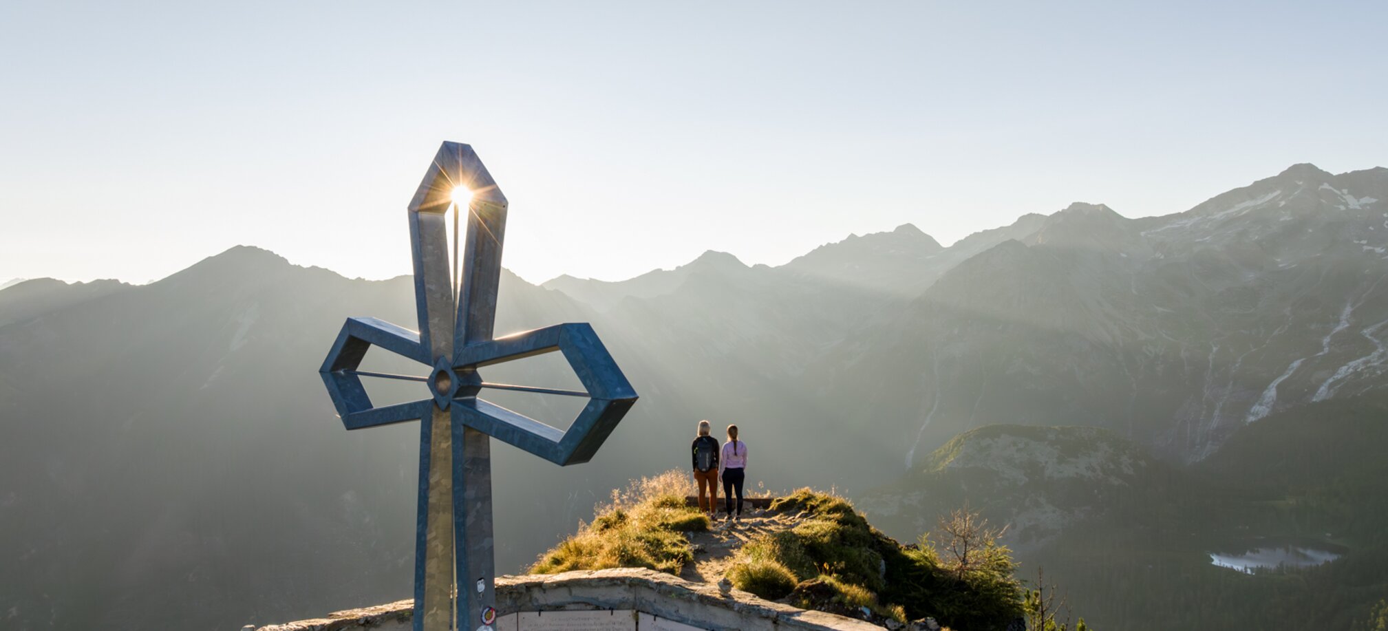 Two women at Graukogel summit cross, sunrise behind mountains, overlooking stunning alpine panorama | © Skigastein, seven frames