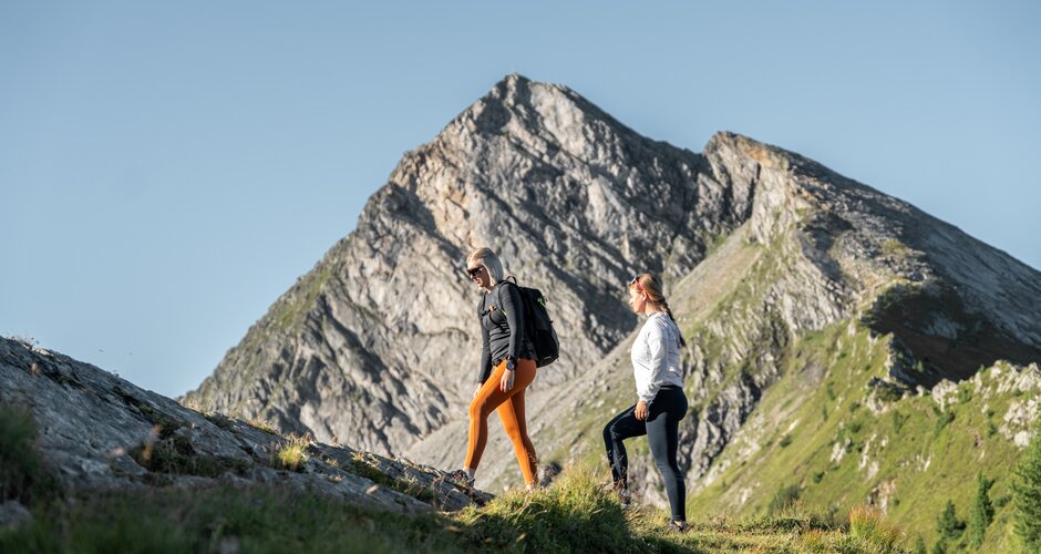 Two women hiking uphill at Graukogel, with a rugged rocky mountain peak in the background under clear blue sky | © Skigastein, seven frames