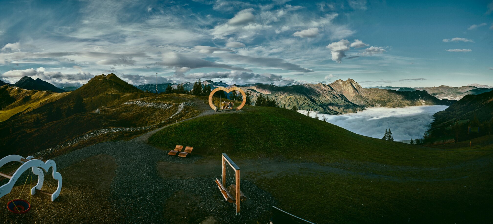 Heart-shaped wooden swing on a hill, mountains and sea of clouds in the valley, dramatic clouds in the sky. | © Großarler Bergbahnen