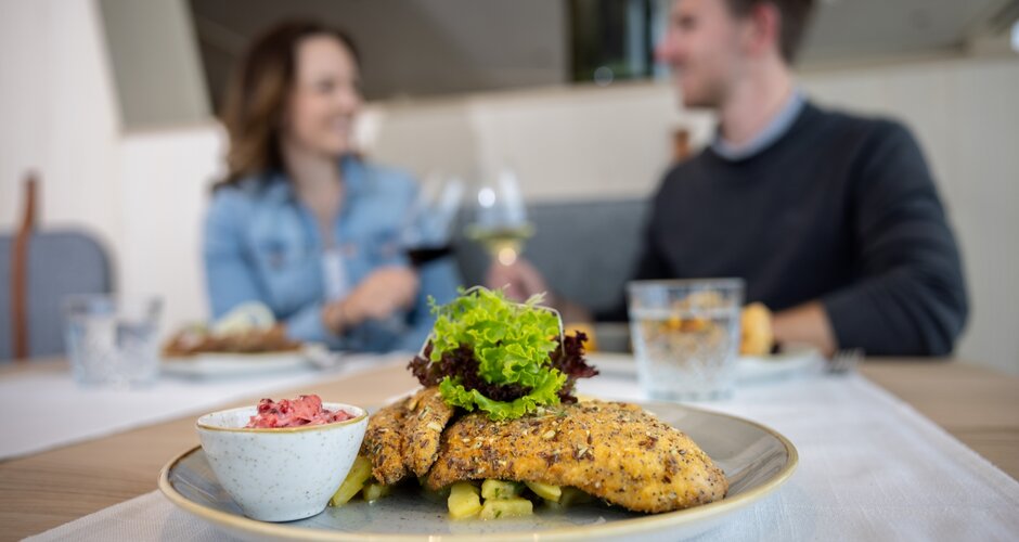 Close-up of a breaded fish fillet with salad and side dish, blurred smiling couple with wine glasses in the background. | © Harald Steiner