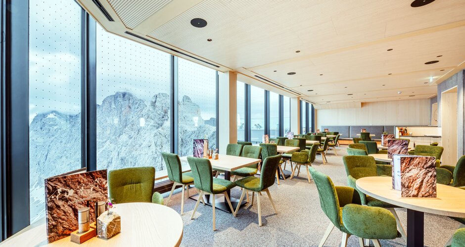 Bright restaurant with large panoramic windows, green upholstered chairs, and wooden tables overlooking the surrounding mountains. | © Christoph Huber