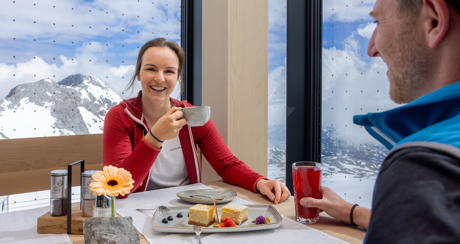 Smiling woman in a red jacket drinks coffee, man with red drink, Alpine panorama through large windows, dessert on table. | © Harald Steiner