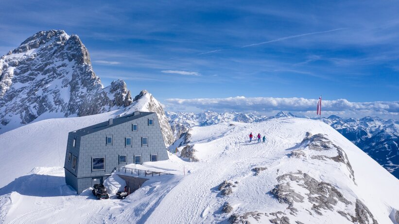 Modern mountain hut on a snow-covered peak with a panoramic view, three people hiking to the summit, Austrian flag in the wind. | © Josh Absenger