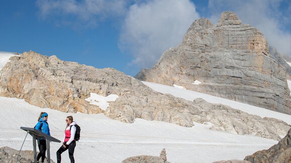 Two women in hiking gear stand at an information board on a glacier, surrounded by rugged rocks and snowfields. | © Birgit Walcher