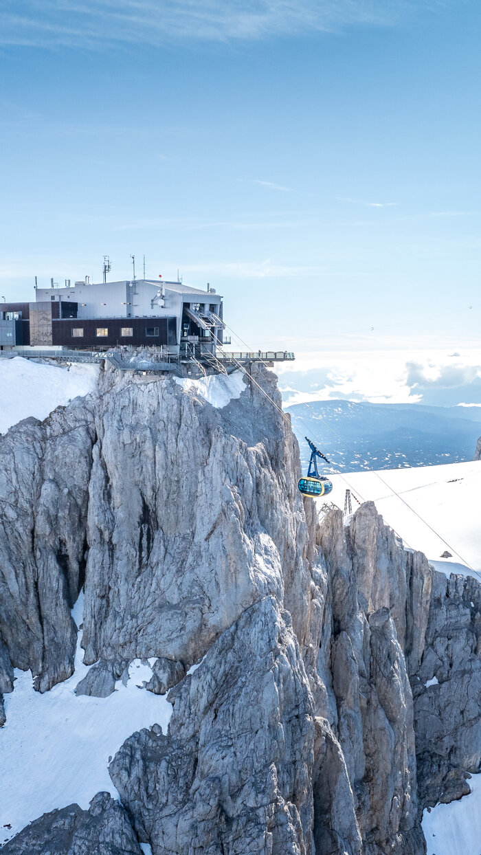 A blue cable car ascends to a modern mountain station on rugged cliffs, offering a wide view of snow-covered peaks and valleys. | © Josh Absenger