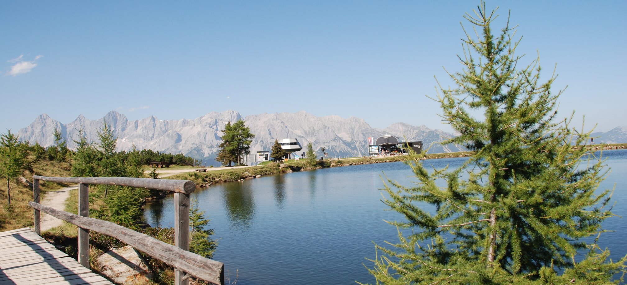 A boardwalk leads along the calm Reiteralm mountain lake, with Dachstein range and two cable car stations in sunny backdrop. | © Reiteralm Bergbahnen