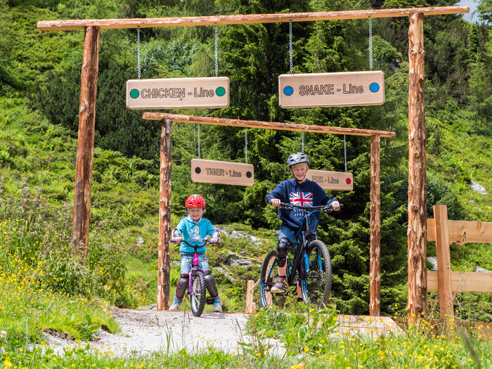 Two kids ride their mountain bikes into signed Junior Trails at Reiteralm, surrounded by wildflowers and green forest trees. | © Reiteralm Bergbahnen