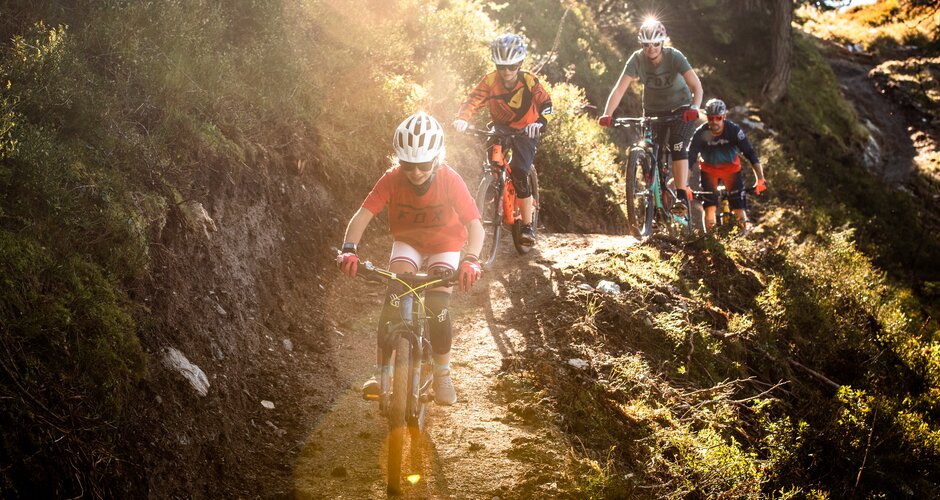 Four mountain bikers, including two kids, ride a narrow forest trail on Reiteralm with warm sunlight filtering through trees. | © Reiteralm Bergbahnen, lorenzmasser.com