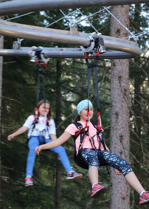 Zwei Kinder hängen gesichert in Gurten und schweben mit der Flyline durch den Wald am Rittisberg. | © Erlebnis Rittisberg