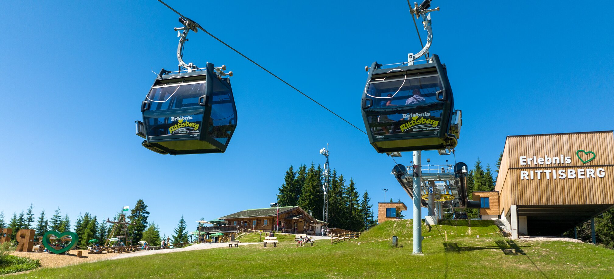 Zwei Gondeln der Rittisbergbahn erreichen die Bergstation, im Hintergrund Spielbereich, Hütten und blauer Himmel im Sommer. | © Rittisberg