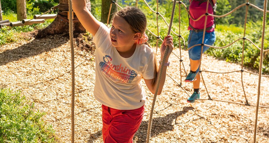 Girl balances on wooden beam in rope park, woman climbs in background. | © Shuttleberg