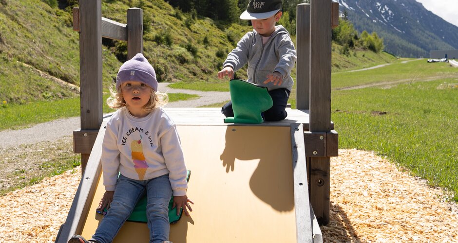 Two small children play on a low wooden slide with green mountains behind. | © Shuttleberg
