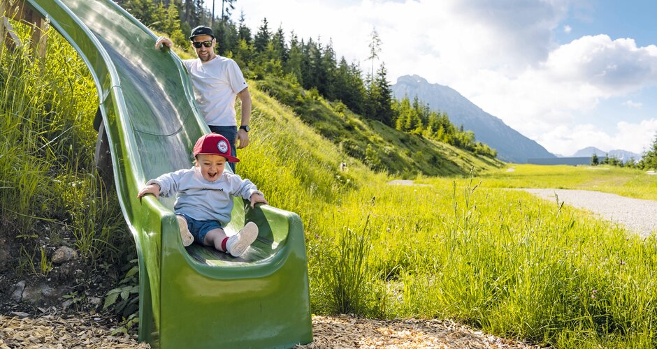 A toddler laughs while sliding down a green slide, an adult stands behind. | © Shuttleberg