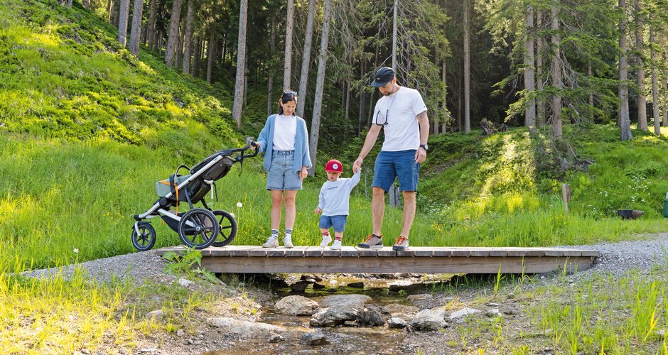 Parents with toddler and stroller cross a small wooden bridge in a green forest. | © Shuttleberg