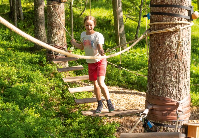 Girl crosses hanging bridge in forest rope course on a sunny day. | © Shuttleberg