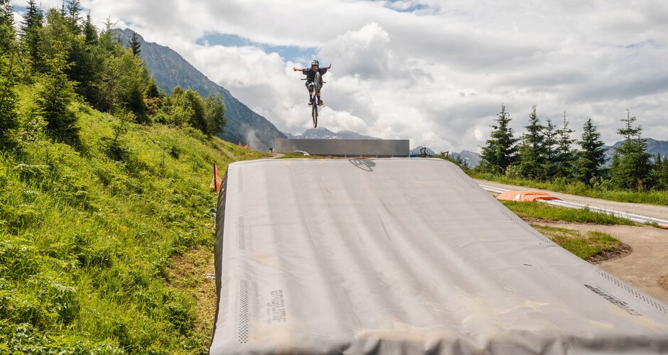 Mountain biker jumps with arms outstretched over Bagjump in green alpine setting. | © Shuttleberg