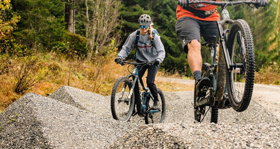 Two mountain bikers ride over gravel hills in the forest, one jumping. | © Shuttleberg