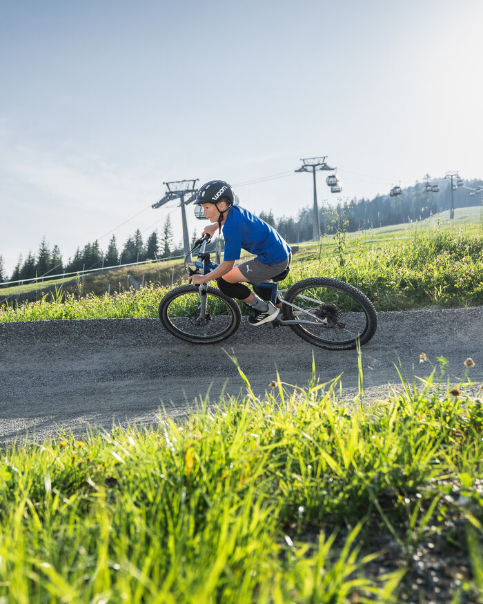A boy wearing a helmet rides a Woom bike on a gravel trail with grassy hills, forest, and cable cars in the background. | © Snow Space Salzburg - Chris Eder