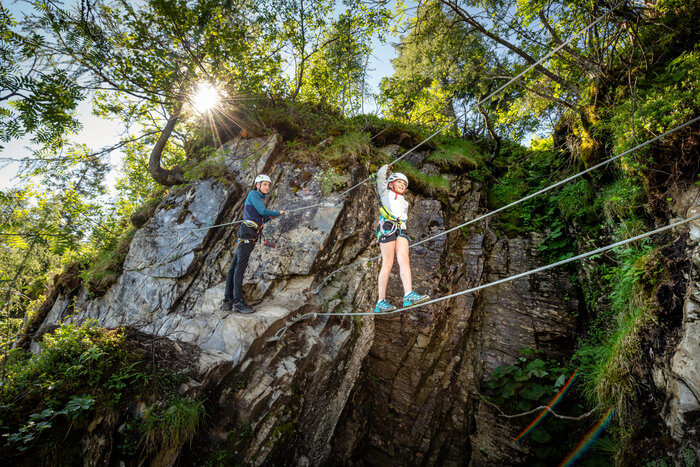 Two kids with helmets and harnesses on rope bridge at rock wall, sunny weather, trees and cliffs in background. | © Snow Space Salzburg, Christoph Huber