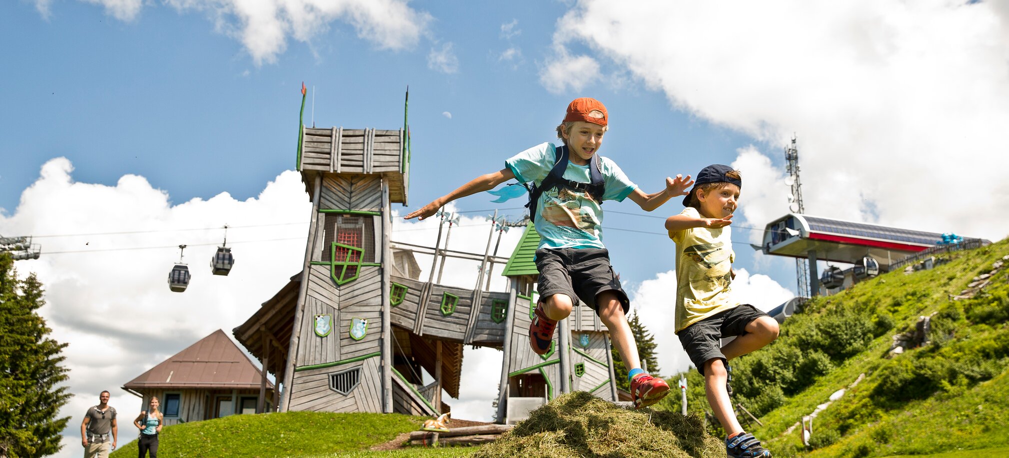 Two kids jump over a grassy mound in front of a fantasy play tower at Geisterberg adventure park in St. Johann, Salzburg | © sanktjohann.com | Mirja Geh