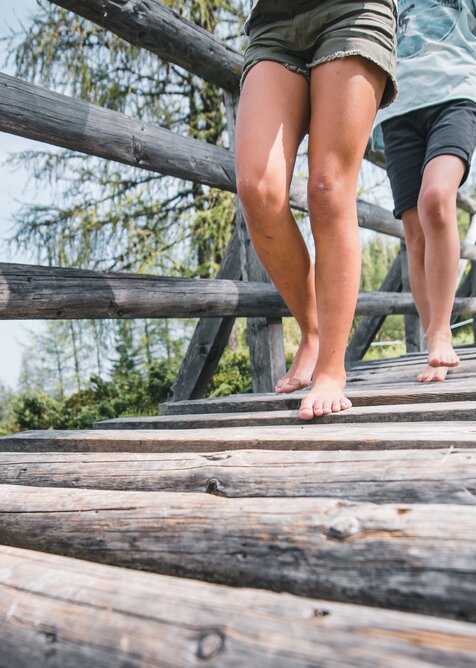 Two people walking barefoot over wooden bridge, close-up of legs, meadow, forest and hikers visible in the background | © Snow Space Salzburg