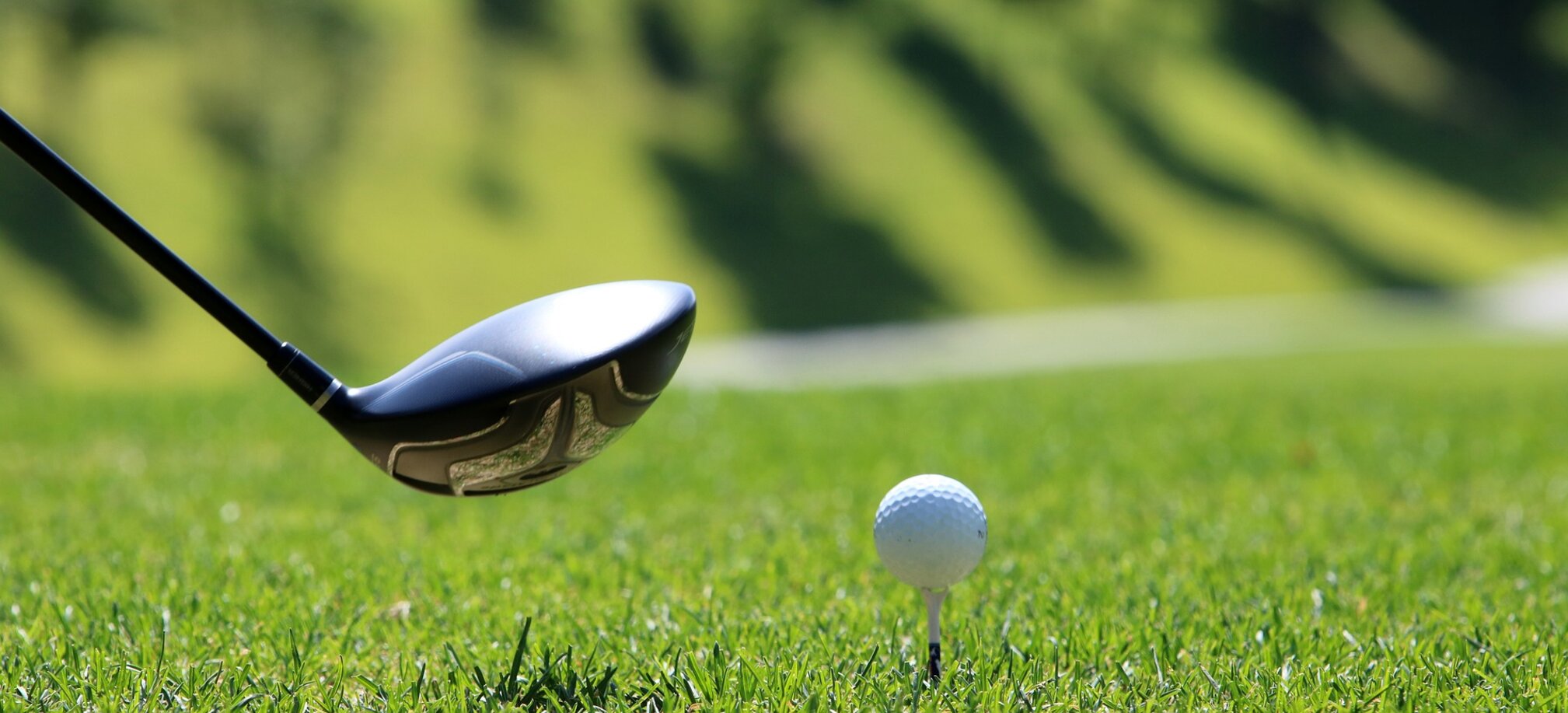 A golf ball on a tee about to be hit with a driver club, surrounded by bright green grass on a sunny fairway.