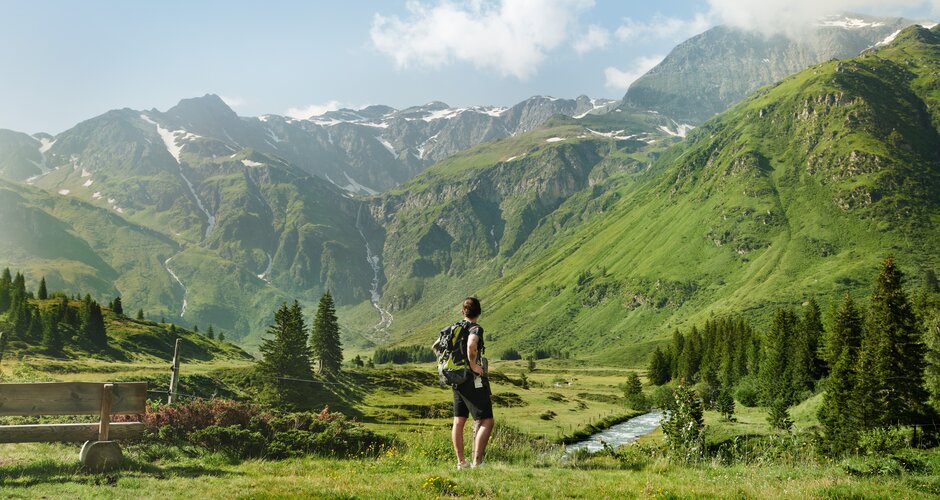A hiker with backpack stands on an alpine meadow in Sportgastein, overlooking a green valley with stream and mountain view | © Gasteiner Tourismus GmbH, Michael Königshofer