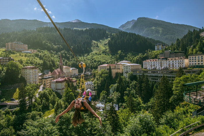 Woman glides on the Flying Waters zipline over rooftops of Bad Gastein, surrounded by forested hills and alpine peaks | © Max Steinbauer Photography